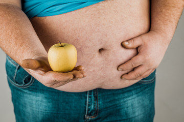 The fat man holds a Apple in front of his bare stomach. The concept of diet and healthy eating