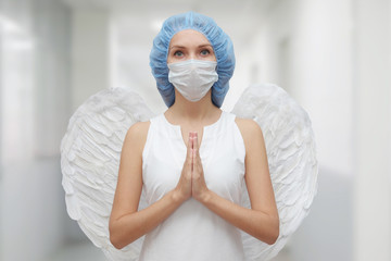 A woman doctor in a medical mask and with white angel wings behind her back prays to God for the...