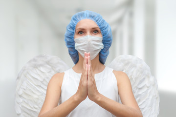 A woman doctor in a medical mask and with white angel wings behind her back prays to God for the...