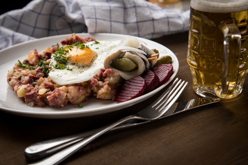 Norddeutscher Hamburger Labskaus mit Rindfleisch Corned Beef, Rote Beete, Kartoffeln, Saure Gurken, Spiegelei, Rollmops und Bier auf Holztisch