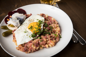 Norddeutscher Hamburger Labskaus mit Rindfleisch Corned Beef, Rote Beete, Kartoffeln, Saure Gurken, Spiegelei, Rollmops und Bier auf Holztisch