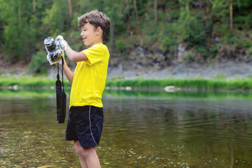 A child is standing in outdoor near the river and forest, holding a camera in his hands and...