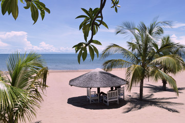 Wooden bungalow surrounded by palms and palm leaves on the beach with sand of light pink color near amazing lake Malawi or Nyasa with turquoise water in Africa. Sunny peaceful african vacation