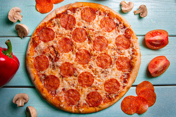 Pepperoni pizza on a turquoise background. Tomatoes, sausage and red pepper on the table.