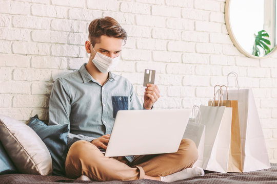 Young Male Shopper In Medical Face Mask Using Laptop On Couch For Online Shopping. Hipster Man In Protective Mask Hold Credit Card In Hand. Digital Payment. E-commerce Store. COVID-19 Home Quarantine