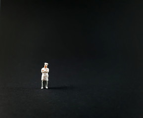 Miniature master chef, standing alone, is pretending to cross his arms against on black background.