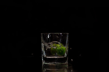 Water splashes out of the garnished glass as a green fresh lemon slice immerses into the water.