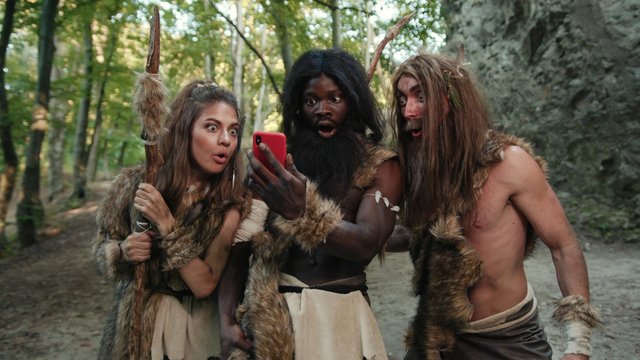 Cheerful Group Of Neanderthal Hunters Using Modern Technology Smartphone Cheering Up Walking Outside Cave In The Jungle. Technology And First People.