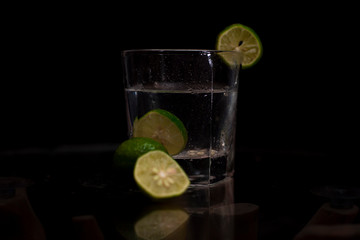 Fresh green lemon slices are immersed into a glass of water and garnished with slices of lemons in a dark copy space background.