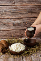Fresh cottage cheese in a wooden bowl with hand pours sour cream from jar on a wooden old background. Rich in Protein and Calcium healthy dairy product, lorw fat soft white cheese. With bread and hay.