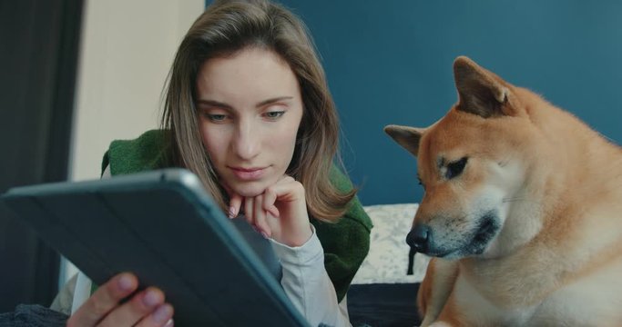 Low Angle View of Young Woman Lying on Bed with Dog Nearby at Home. She Holds Tablet, Looks at Its Screen and Strokes Her Dog. Animal-Human Friendship Concept. Slow Motion Shot