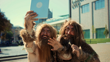 Stone-age wild men with long hair animal skin using digital tablet taking selfies rejoicing with fun like modern people. Neanderthals in civilization.