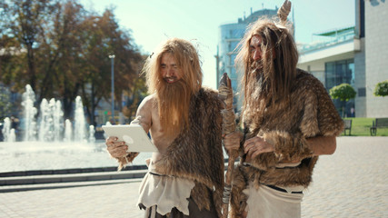 Backward primitive neanderthals using tablet pc in the street. Two aggressive cavemen in animal fur growling exploring technology and modern life.