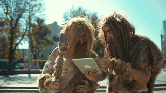 Primitive neanderthals using tablet pc computer like modern people. Ancient huntsmen exploring present life and civilization. Evolution degradation.
