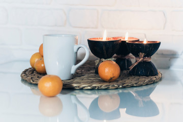 Burning spa aroma candles in coconut shell, cup of tea, tangerines on wicker stand on a glass white table, cozy home interior background.