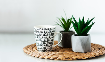 Cozy home interior decor: white and black cup and ornamental plants in pots on a wicker stand on a white table in the room. The quarantine concept of stay home.