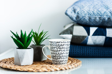 Cozy home interior decor: white and black cup and ornamental plants in pots on a wicker stand on a white table in the room. Blue pillows on background. The quarantine concept of stay home.