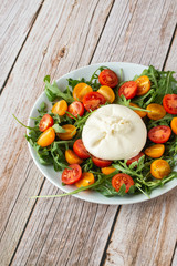Tasty, fresh salad with mozzarella burrata, arugula, cherry tomatoes and basil on a white plate on a wooden background.Close up. Healthy Italian food. Selective focus