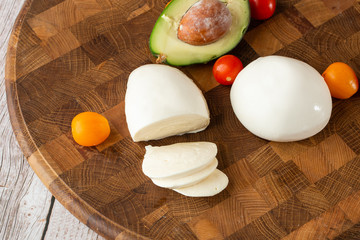 Overhead photo of buffalo mozzarella cheese with cherry tomatoes, basil leaves, avocado, ingredients of salad, pesto sauce, olive oil, on rustic wooden cutting board with copy space. Italian cheese