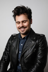 Obraz premium Funny bearded man wearing biker jacket, smiles strangely and looks at the camera with closed eyes. Vertical shot.