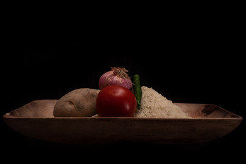 A pile of rice, potatoes. fresh onion, tomato and green chilli are kept on a wooden tray in a dark copy space background.