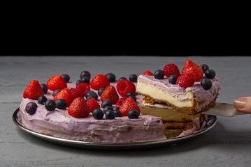 Woman divided cheesecake and take one slice to offer it. Home made Cheesecake with Strawberry, blueberry and raspberry on grey wooden background.