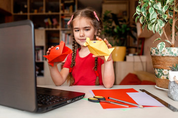 Girl doing origami fish with color paper looking video on laptop, online workshop and distant...