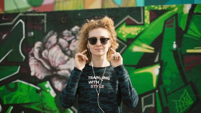 red-haired girl in sunglasses puts on headphones on a background of graffiti