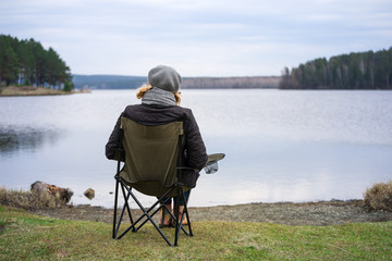 An adult woman enjoys nature, sitting in a camp chair on the Bank of a forest pond in early spring. The concept of a healthy lifestyle for older people.