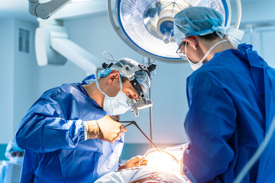 Process Of Surgery Operation Using Medical Equipment. Two Surgeons In Operating Room With Surgery Equipment And In Binocular Glasses. Medical Background.