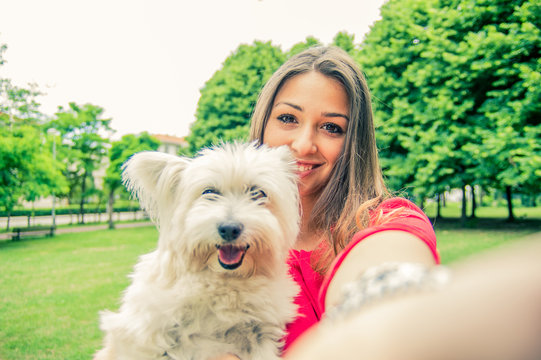 Beautiful young woman having fun taking a selfie portrait with her white dog outdoor at the park. Love between people and animal concept