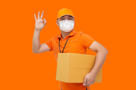 Delivery man holding parcel box and wearing protective mask for prevent virus Covid-19 and showing ok isolated on orange background, Online shopping shipment and fast express delivery service concept