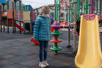 Blond hair girl, wearing a medical mask, closed playground on the background. A child in a protective mask. Warning of the dangers and safety quarantine measures against COVID-19. Coronavirus pandemic