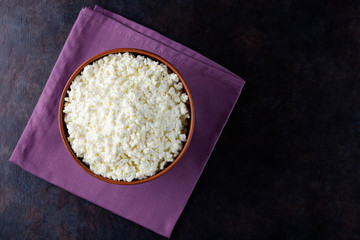 Cottage cheese on a dark background. Fresh cottage cheese in a bowl. Clay plate with soft cheese on a linen napkin. Minimalism. Top view. Copy space