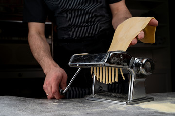 Chef in an apron is cutting raw dough in a pasta machine cutter, male hands visible, close up