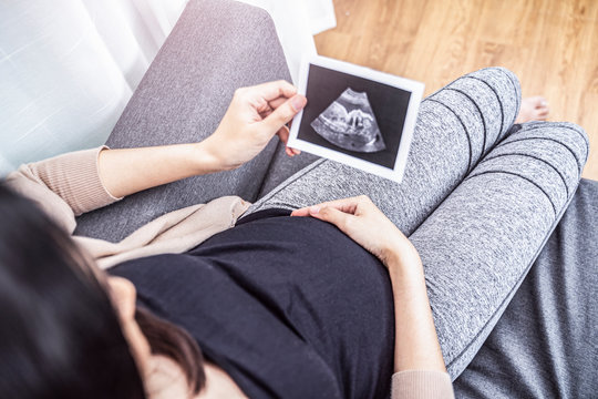 close up asian pregnant woman sitting on sofa holding ultrasound baby picture while placing hand on belly, resting relaxing in living room from hormone stress, wearing comfy stretch pants and cardigan