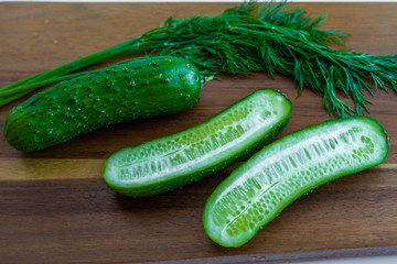 Two halves of cucumber and one whole cucumber are laying on a wooden cutting board together with fresh green dill, closeup macro view