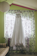 A white wedding dress is hanging on a hanger against a white window with curtains in the bedroom. Morning and preparation of the bride. Photography, concept.