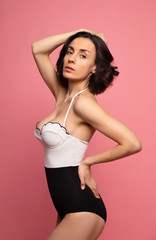 Fototapeta premium Portrait of a gorgeous trendy fit woman in swimwear while she enjoying rest with sunlight isolated over pink background. Travel, tourism, journey, trip, tour, summer concept