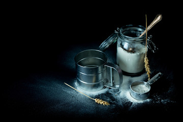 Metal flour sifter, jar and measuring cup on black background