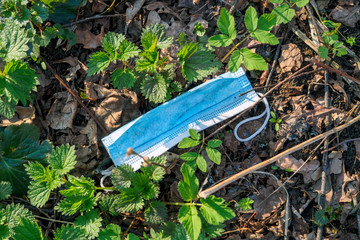 Blue disposable medical face mask between green plants and old dry leaves, thrown away like garbage on nature