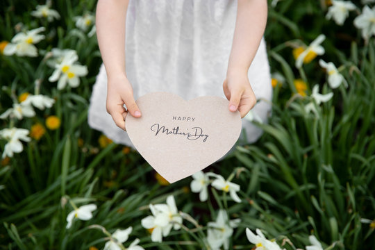 Happy Mother's Day written on a paper card held by a girl on a flower and grass background