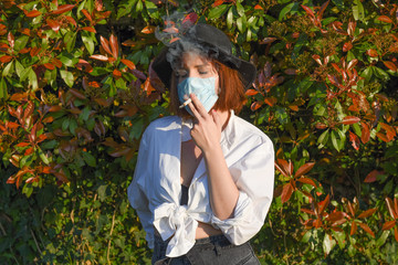 Girl with surgical face protection mask on the natural background smoking a cigarette.