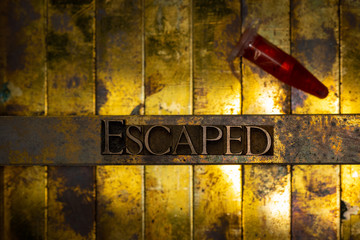 Photo of real authentic typeset letters forming Escaped text with red fluid filled laboratory vial on vintage textured grunge copper and gold background