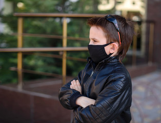 Little boy stays outdoors wearing a black hygyenic medical mask,.frowns and folds his hands. Cancelled vacation, bad mood due to coronavirus covid-19