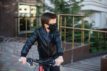 Fototapeta na wymiar Boy in a leather jacket wearing a black hygyenic medical mask outdoors in the yard in black sunglasses, riding a bike. View from the side. Cancelled vacation, bad mood due to coronavirus covid-19
