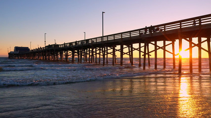Fototapeta premium People walking on the Newport Beach pier during sunset as waves slowly roll in to the beach.
