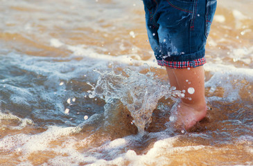 A kid, little boy, staying in leans shorts in water on the beach. Sea foam is coverig his feets. Close up splash of sea water. Summer concept, happy vacation, carefree childhood. Space for text.
