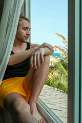 young bearded guy is resting sitting on the windowsill and looking at the sea