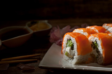 Sushi on a white plate. Sushi roll with sauce and spices on a black background. Food on a wooden table with dark boards. Sakura color on a plate. Flower with rose petals. Place for text near land.